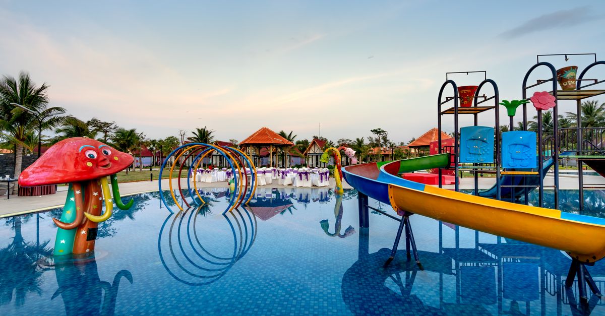 Colorful waterpark with slides, mushroom-shaped feature, and palm trees reflecting in pool.