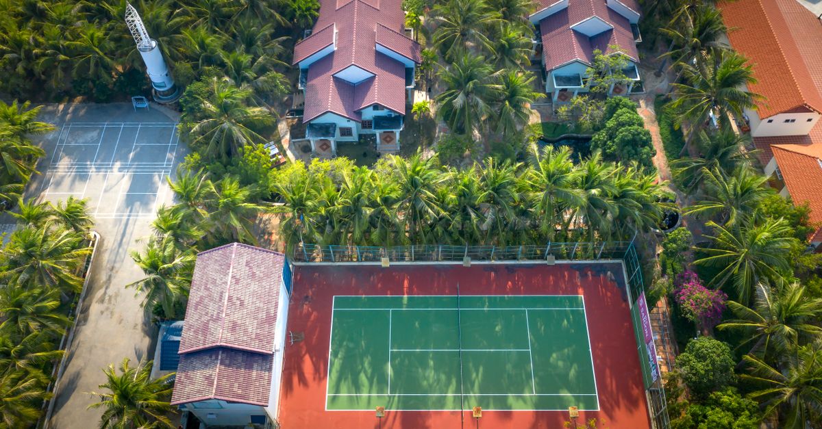 Discover a vibrant tennis court nestled amid lush palm trees and tropical residences from a stunning aerial perspective.