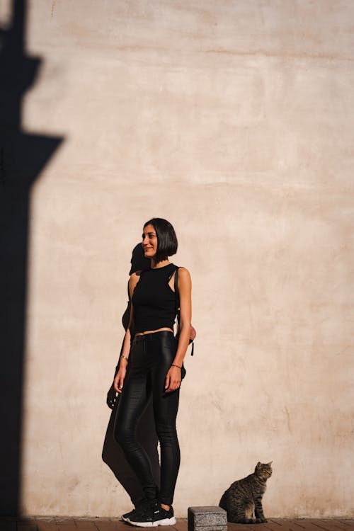 Woman in Black Clothing and a Cat Standing Beside a Concrete Wall