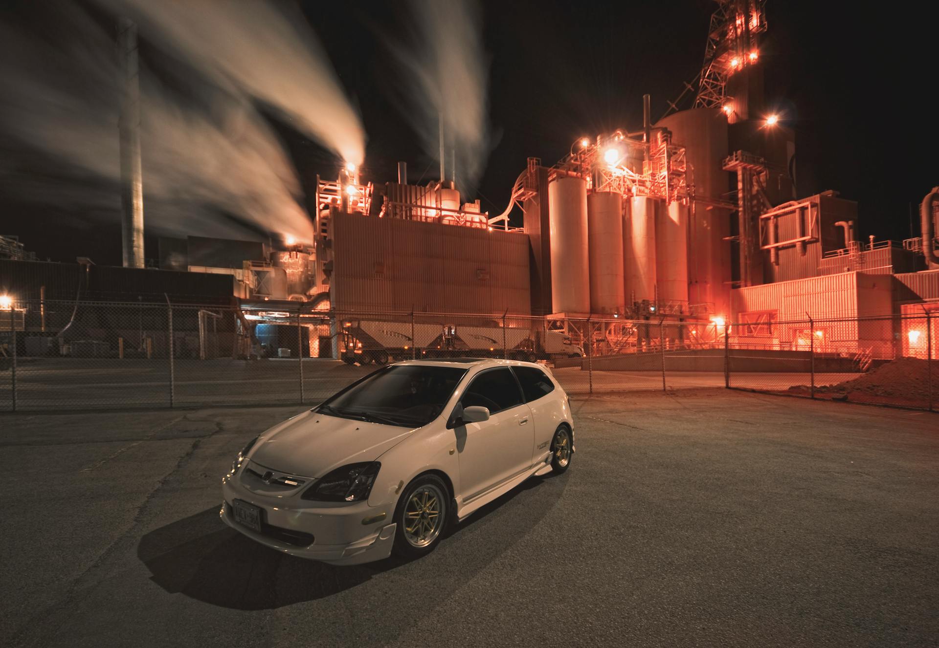 A white car parked in front of an illuminated industrial plant at night with smoke rising.