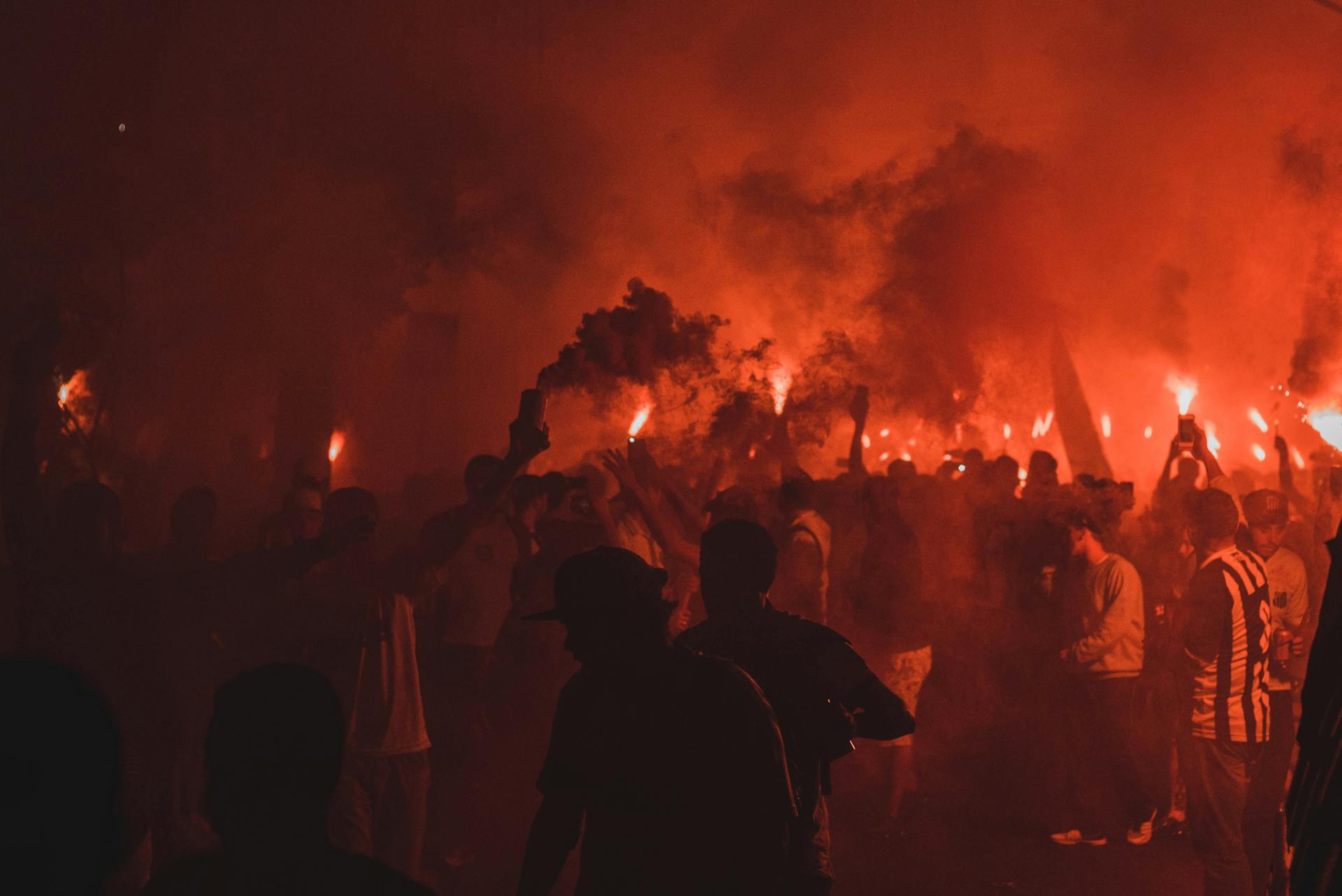 An energetic night celebration with people holding flares amidst smoke and vibrant flames.
