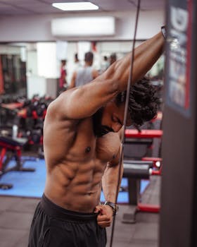 Shirtless muscular man stretching arms in a gym setting, focusing on fitness.