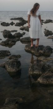 A woman in a flowing white dress stands on rocks by the sea during a calm day.
