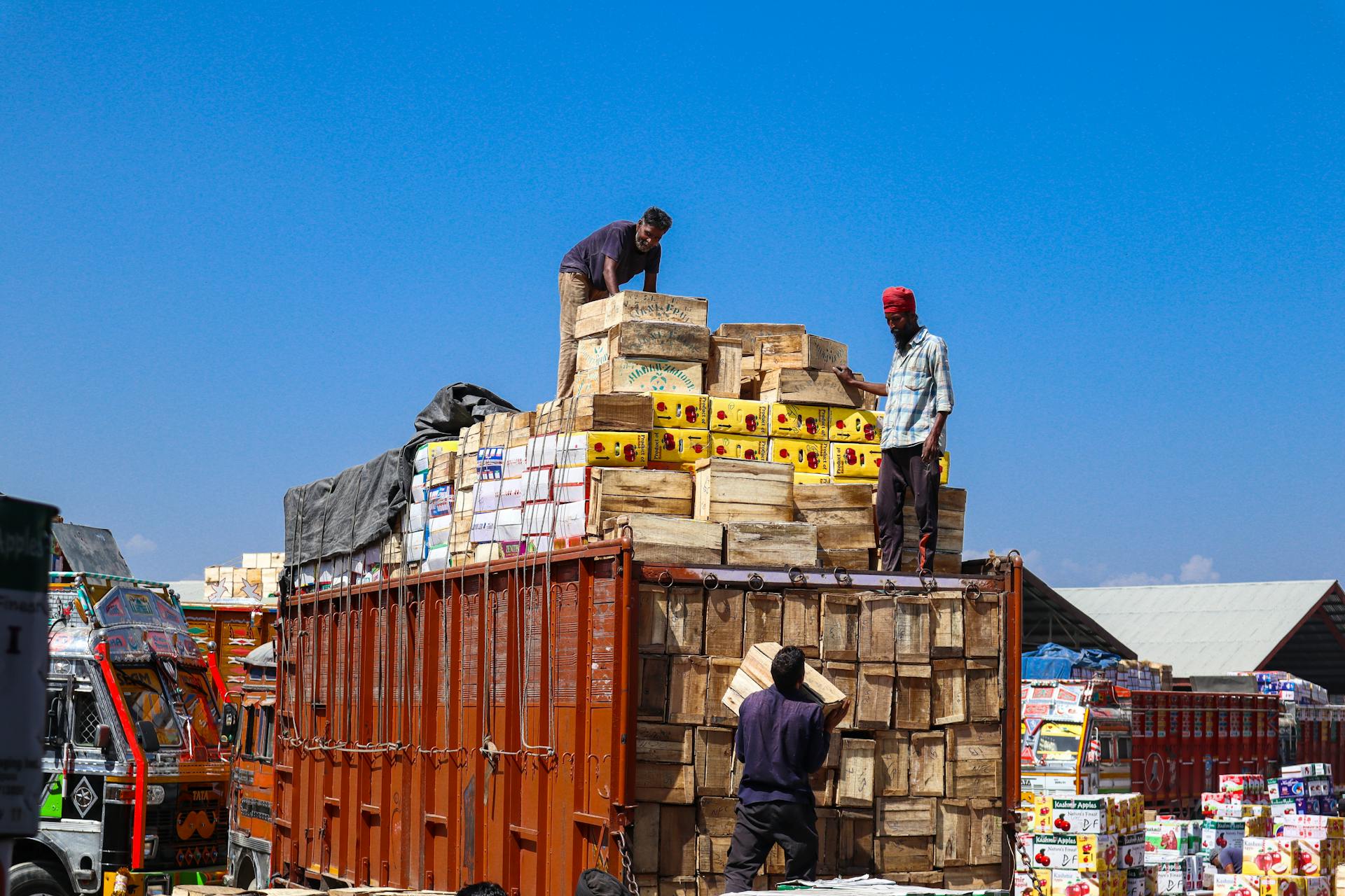 Three men loading wooden boxes on a truck under a clear blue sky in Sopore.