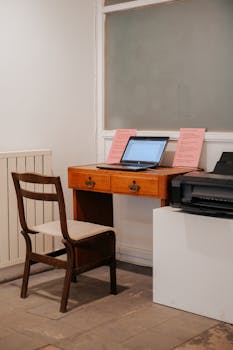 A minimalist workspace featuring a wooden chair and table with a laptop and printer.