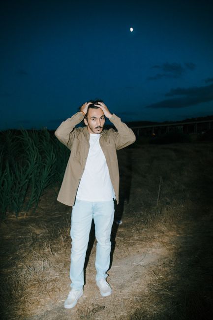 A man with a mustache standing outdoors at night under a moonlit sky, hands on head.