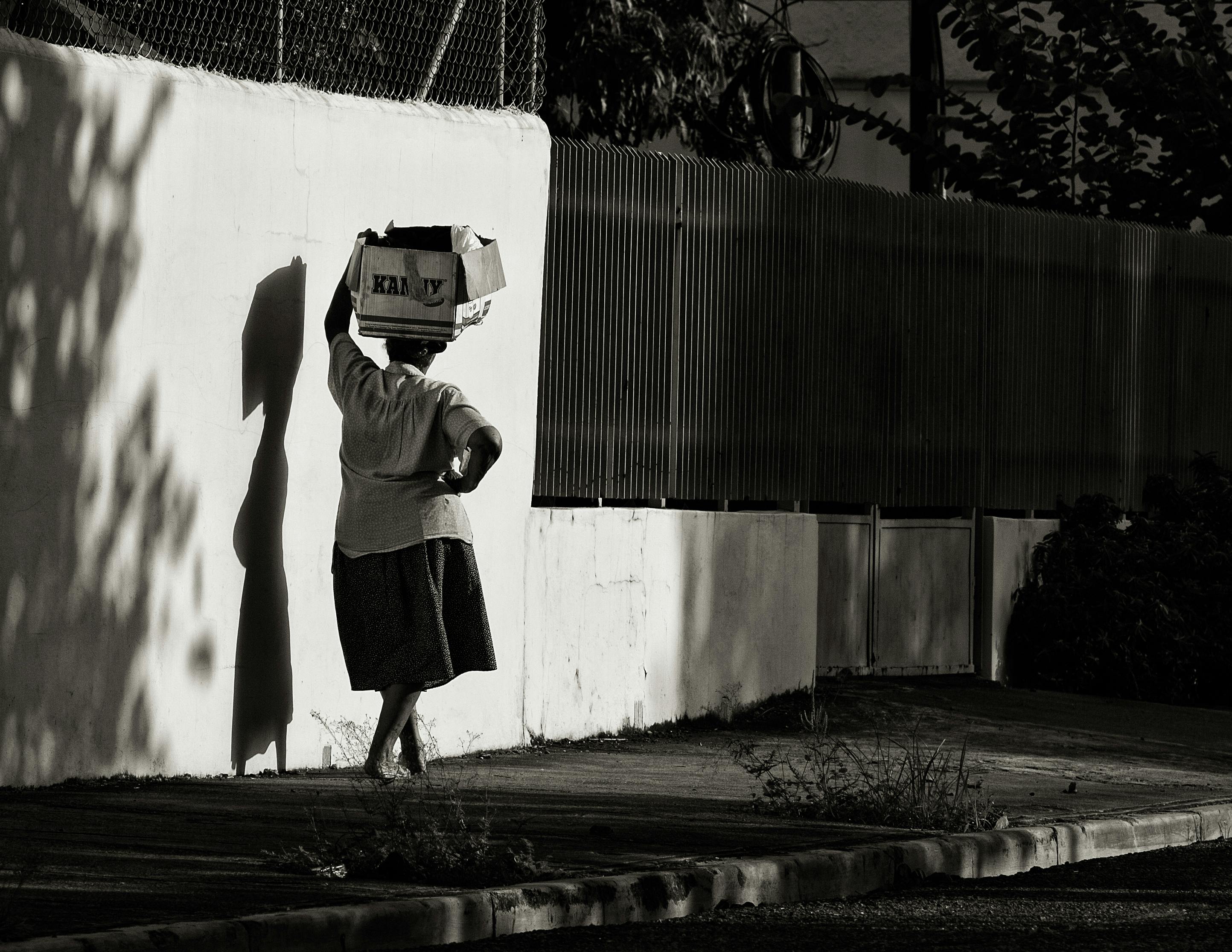 Black and white photo of an elderly woman carrying a box on a city sidewalk.