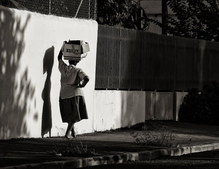 Black And White Picture Of Woman Walking On A Sidewalk And Carrying A Cardboard Box On Her Head 