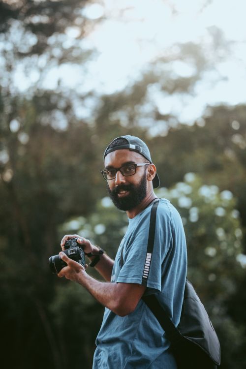 Free Bearded man in a blue shirt holding a camera outdoors in a natural setting. Stock Photo