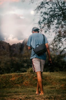A man walks through grassy terrain carrying a bag and camera during twilight. Captured from behind.