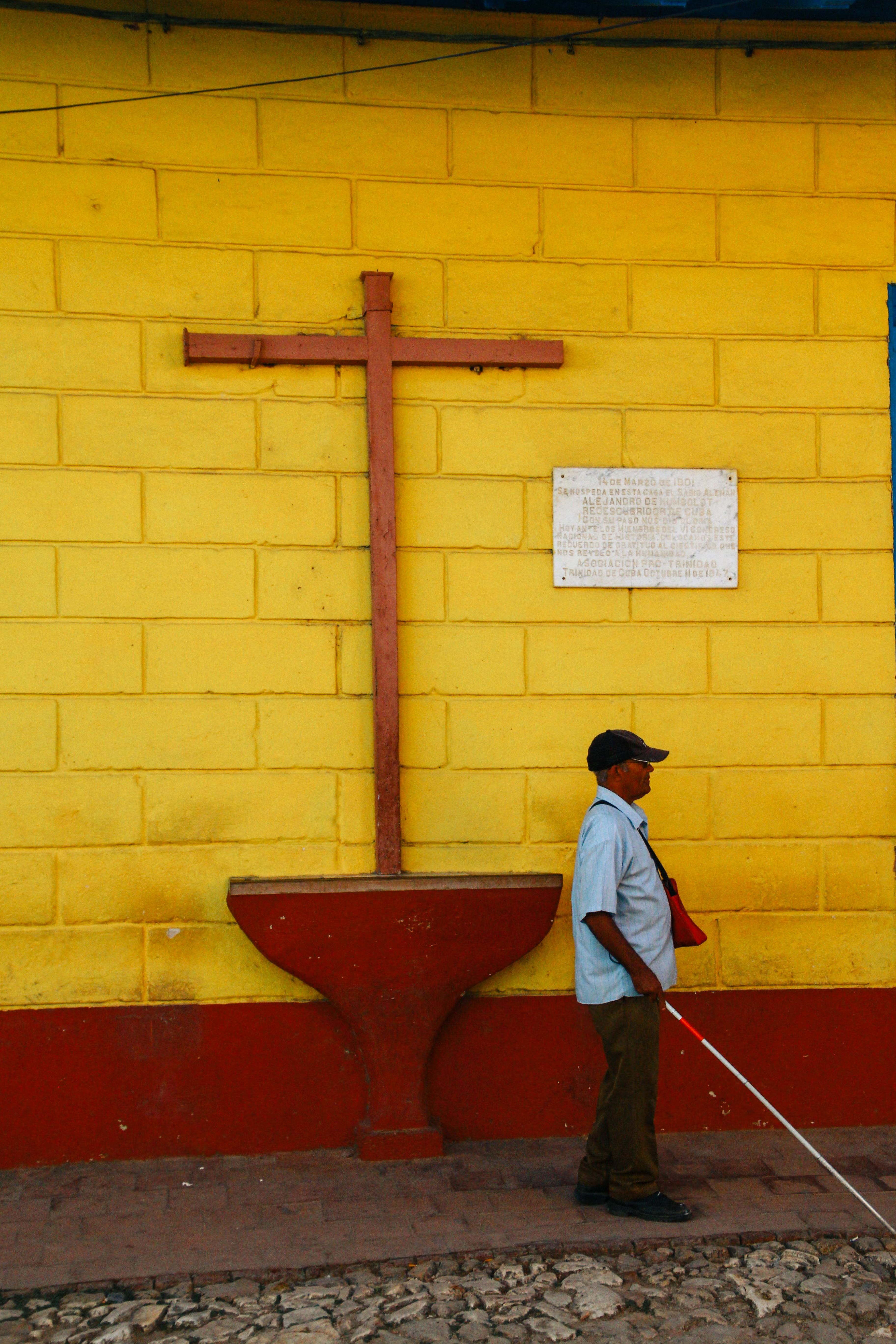 A man with a walking cane passing by a vibrant yellow wall with a cross.
