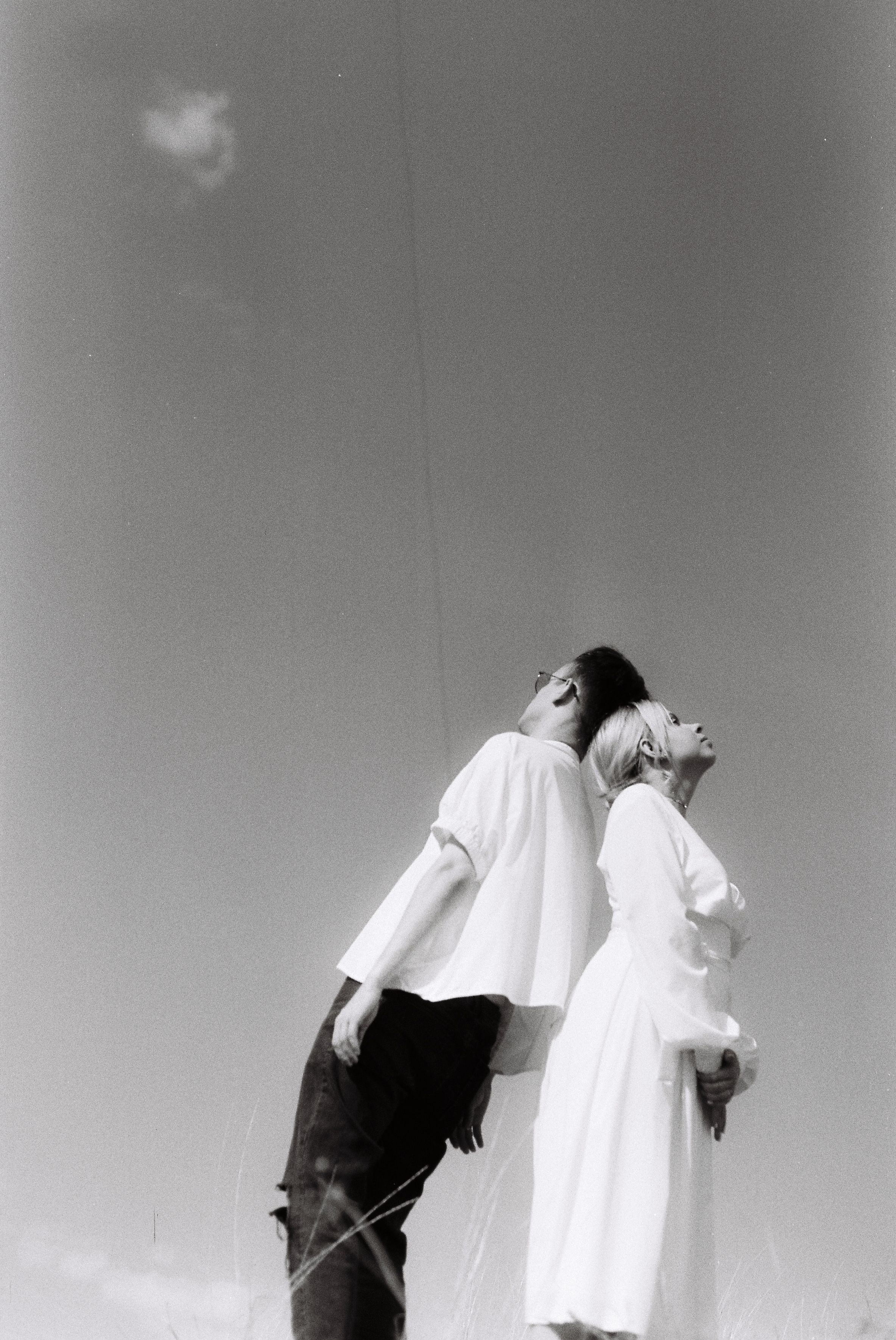 Black and white image of a couple leaning back to back under the open sky.