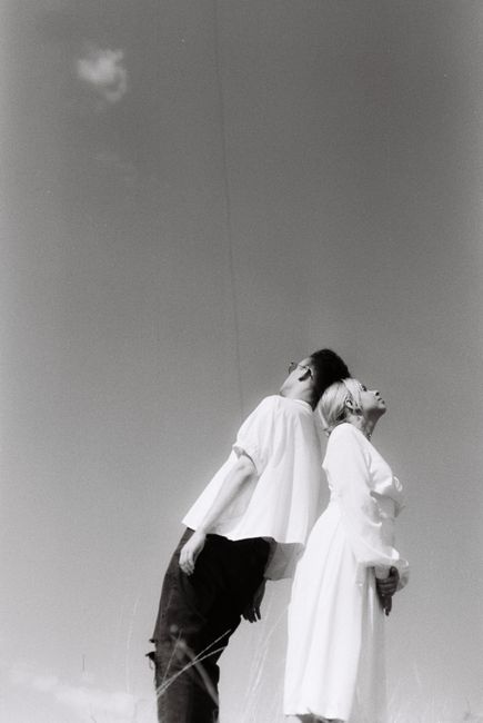 Black and white image of a couple leaning back to back under the open sky.