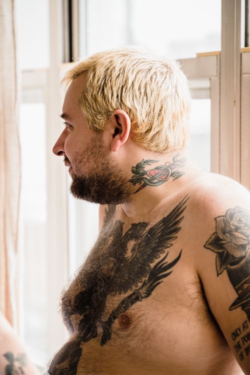 Free A plus-sized topless man with tattoos and blonde hair stands by a bright window. Stock Photo
