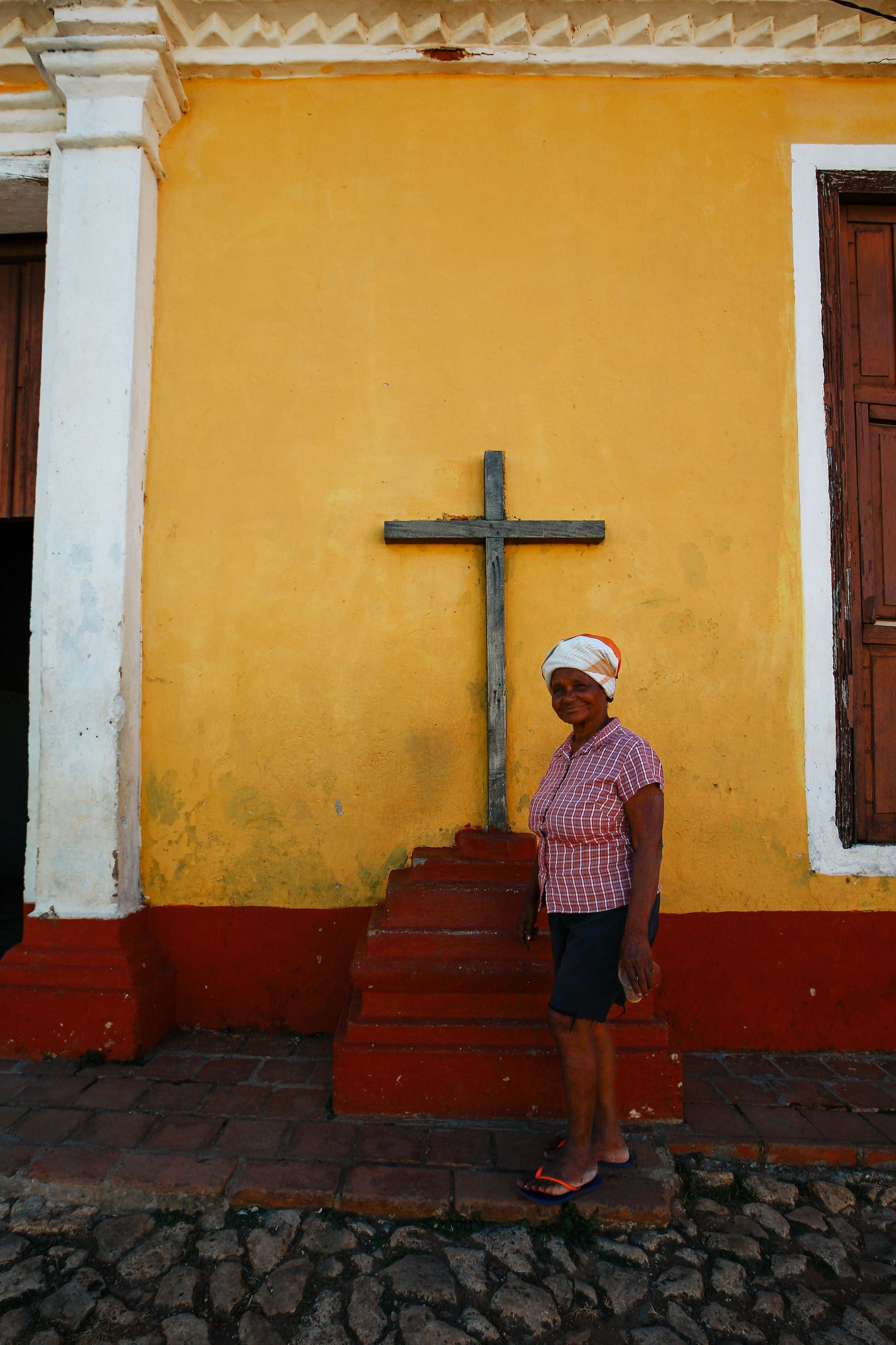 A woman standing in front of a cross on a yellow wall, smiling outdoors.