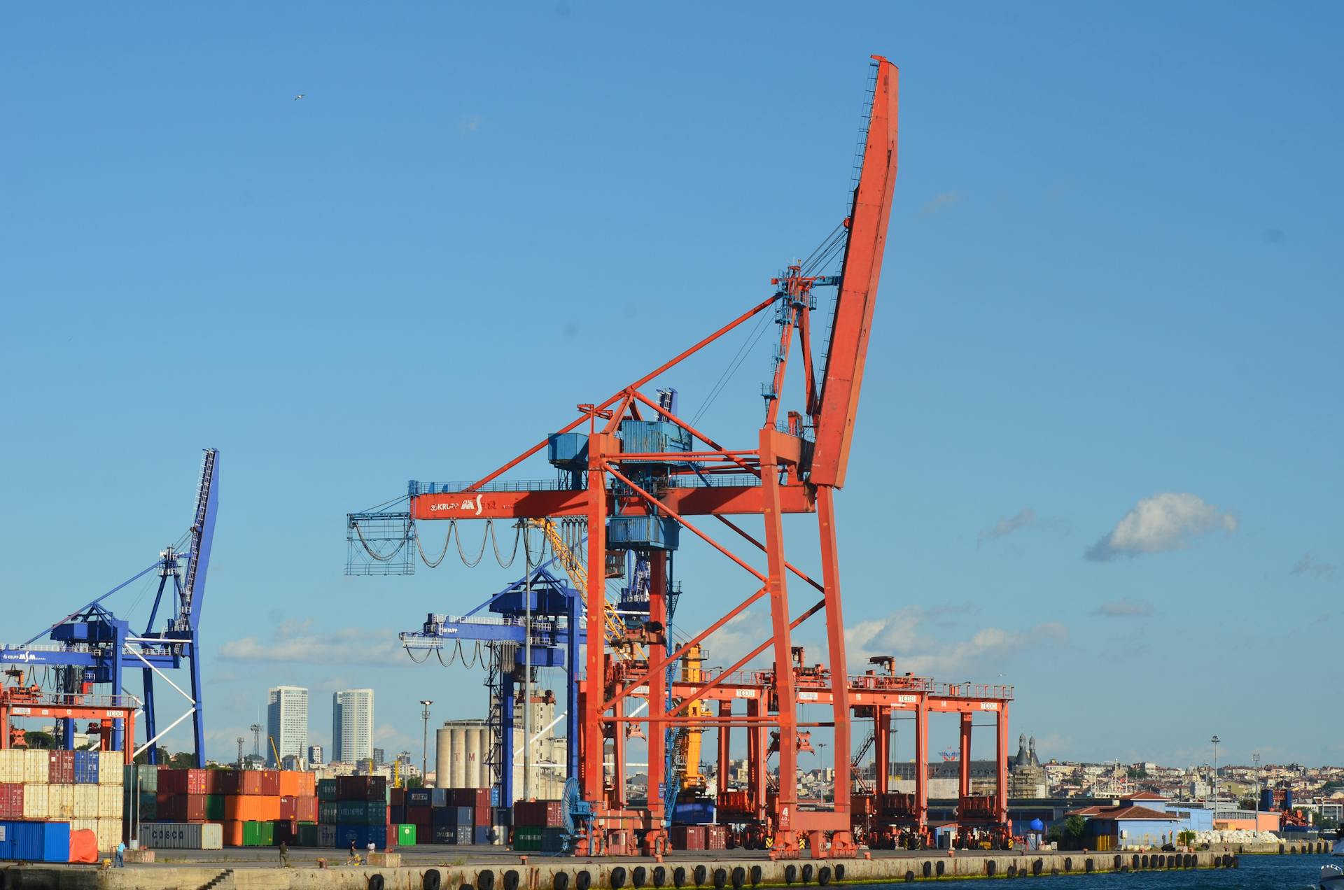 A bustling harbor with large cranes and stacked shipping containers under clear blue skies.