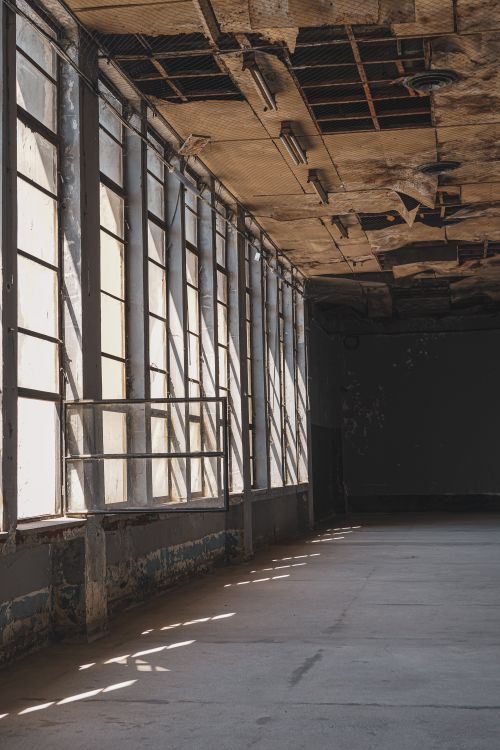 Free A spacious, abandoned industrial interior with sunlight streaming through tall windows casting strong shadows. Stock Photo