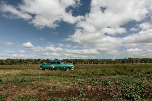 A classic green car parked in a scenic grassy field with a cloudy sky backdrop.