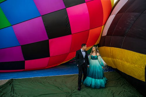 Man in Gray Suit and a Woman in Blue Green Gown Standing Together Beside Air Balloons on Ground