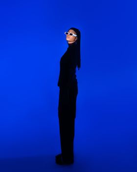 A fashionable woman stands against a vibrant blue background, wearing trendy black attire and sunglasses.