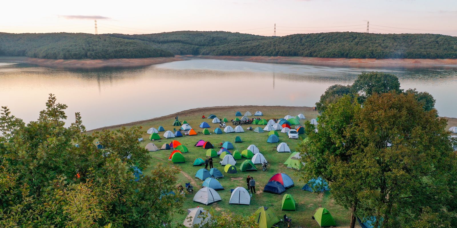 Colorful tents set up beside a tranquil lake amid greenery at sunset