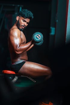A shirtless man engages in an intense workout with dumbbells in a gym setting.