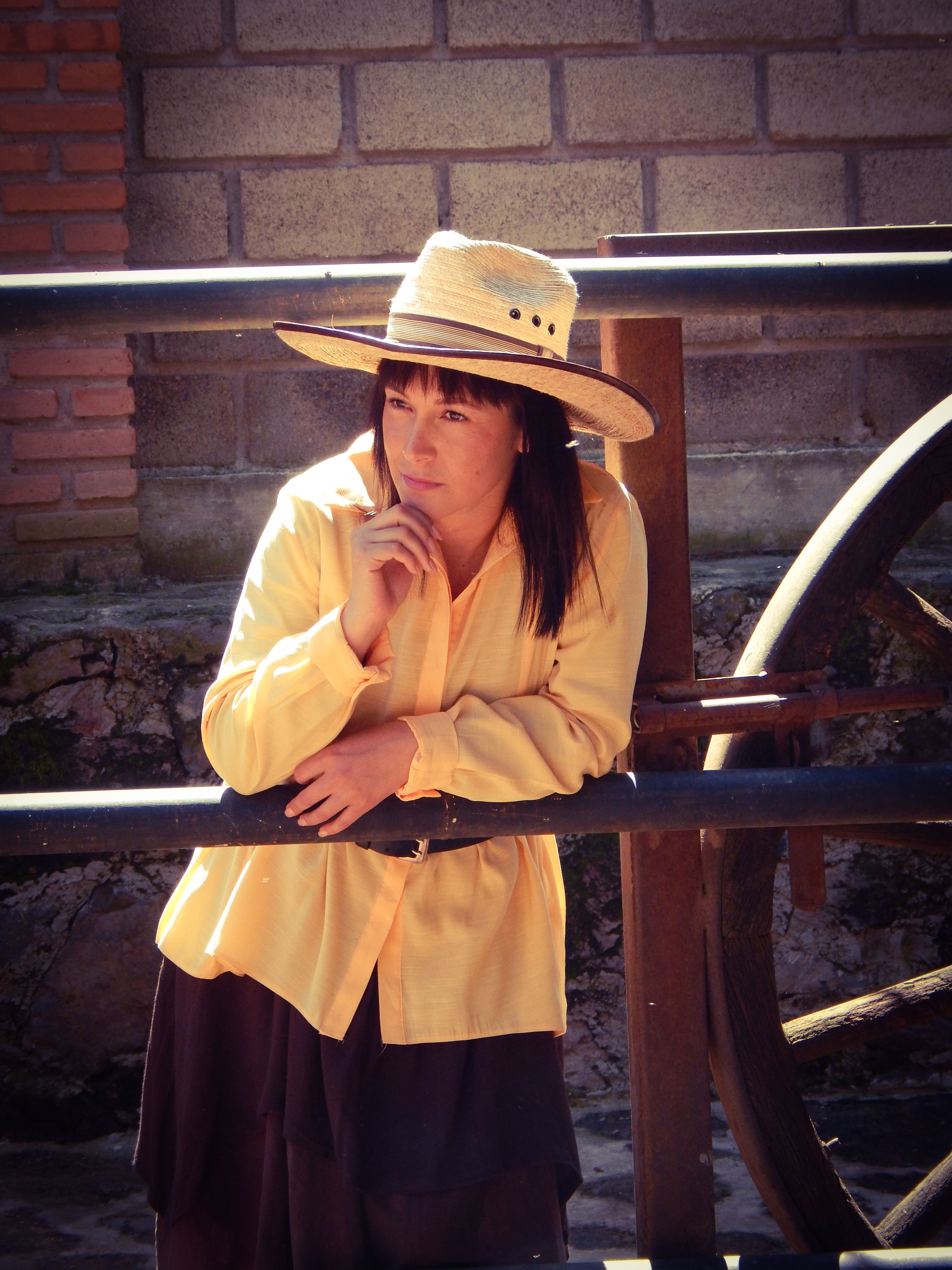A woman in a yellow jacket and hat poses pensively by a historic wheel in San Salvador Cuauhtenco.