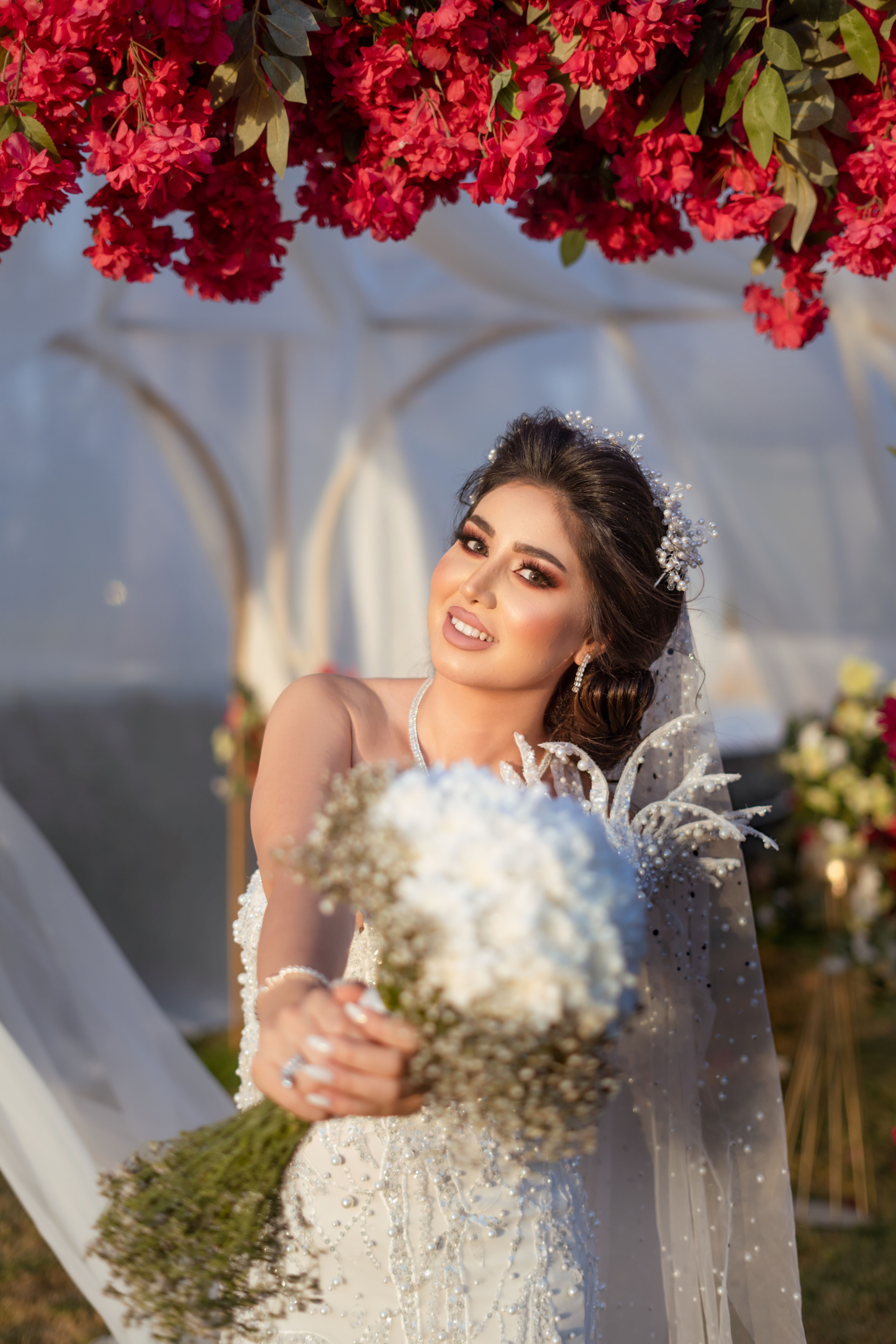A beautiful bride in a wedding gown holds a bouquet under a vibrant flower arch outdoors.