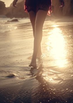 A woman walks barefoot by the seashore reflecting golden sunlight at sunset.