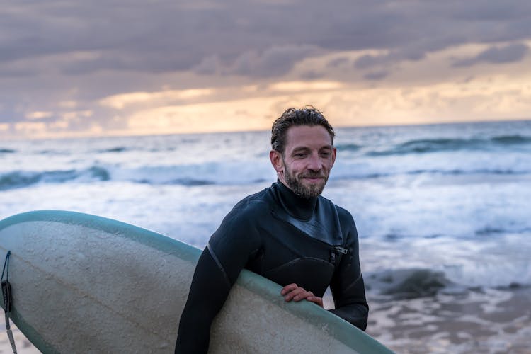 Bearded Man In Black Rash Guard Holding A Surfboard