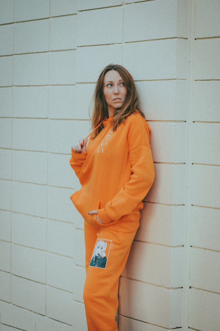 Woman Wearing Orange Hoodie Sweater And Jogger Pants Leaning On The Wall 