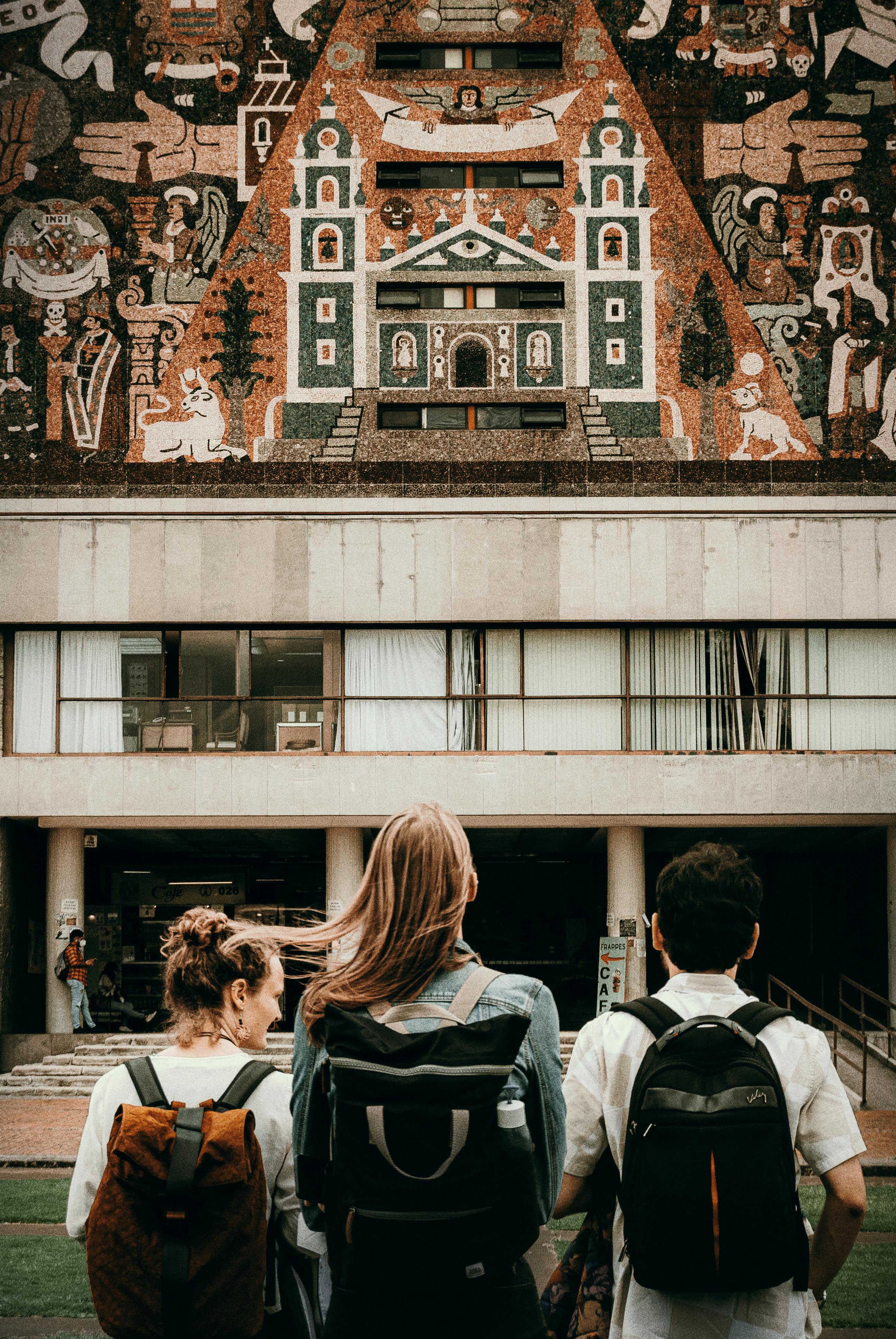 Three young tourists with backpacks admire an elaborate building mural. Urban exploration.