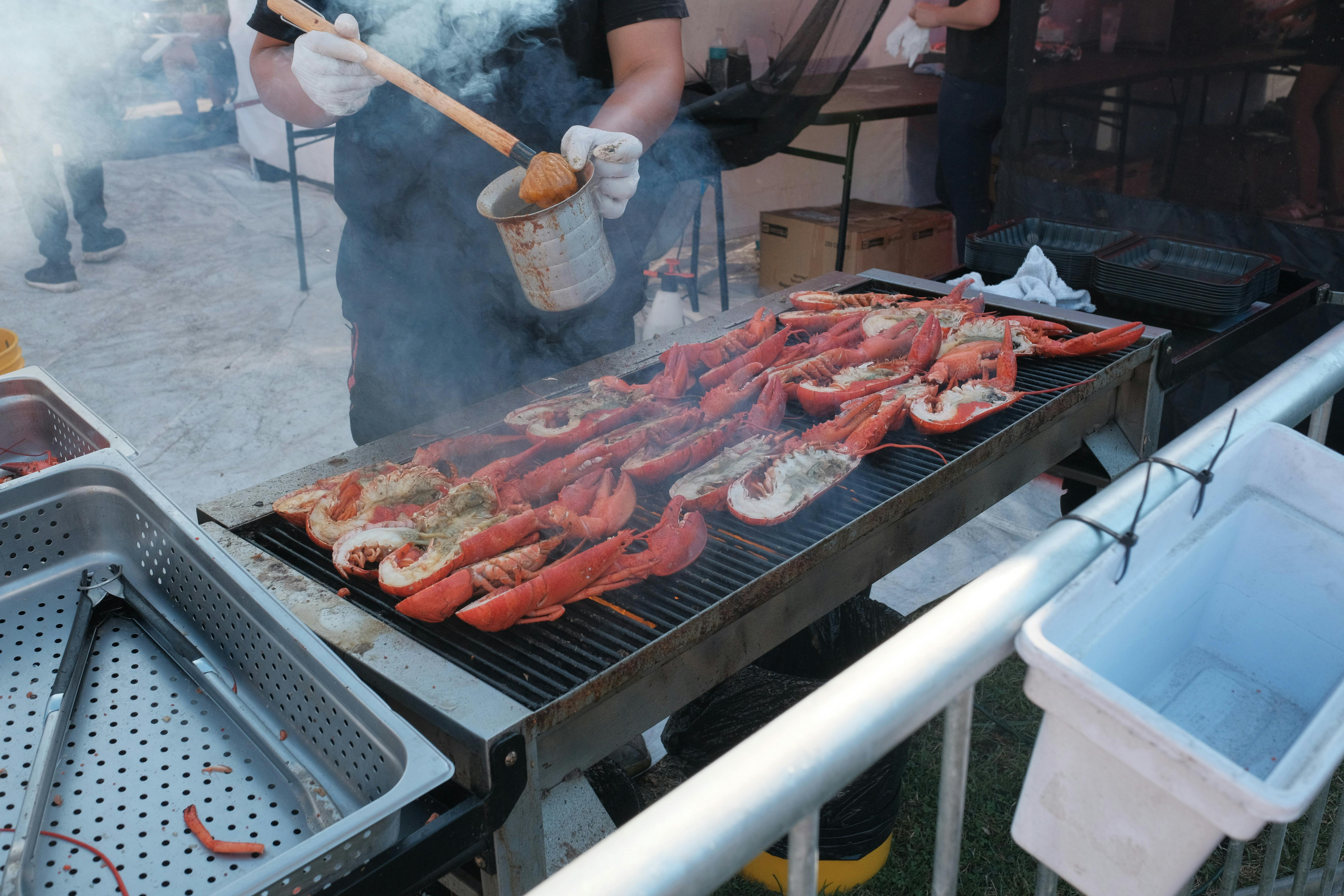 Lobsters being grilled by a man at an outdoor barbecue event with smoke rising.