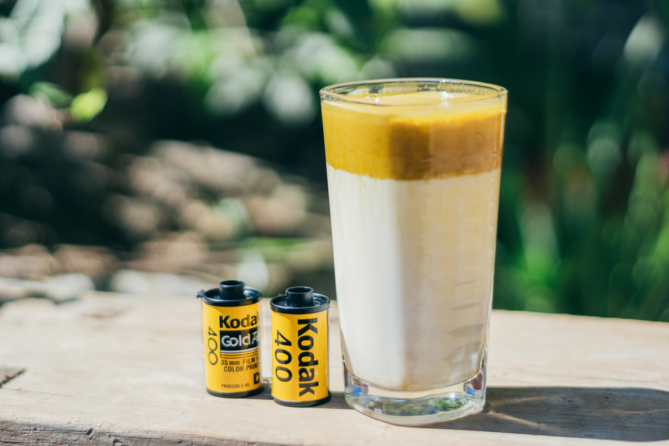 A glass of refreshing layered juice beside Kodak film rolls placed on a wooden table outdoors.