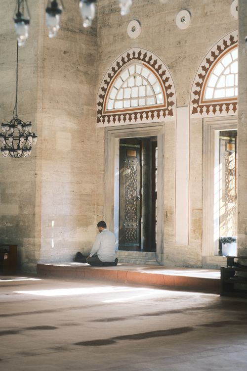 Free A person prays inside a beautifully lit mosque with ornate windows and chandeliers. Stock Photo
