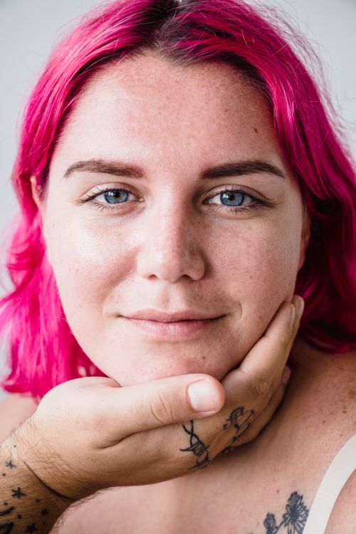 Free Intimate close-up portrait of a woman with vibrant pink hair and a tattooed hand touching her face. Stock Photo