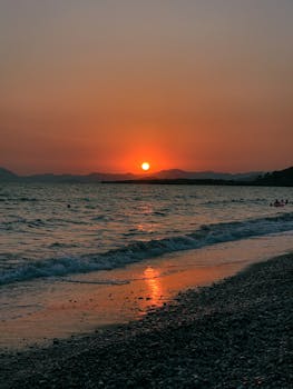 A calming beach sunset with waves, silhouetted mountains, and an orange sky reflected on the water.