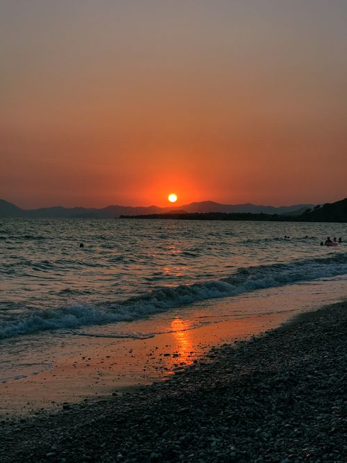 Free A calming beach sunset with waves, silhouetted mountains, and an orange sky reflected on the water. Stock Photo