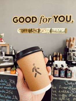 A hand holds a paper coffee cup in a chic cafe with signage reading 'GOOD for YOU.'