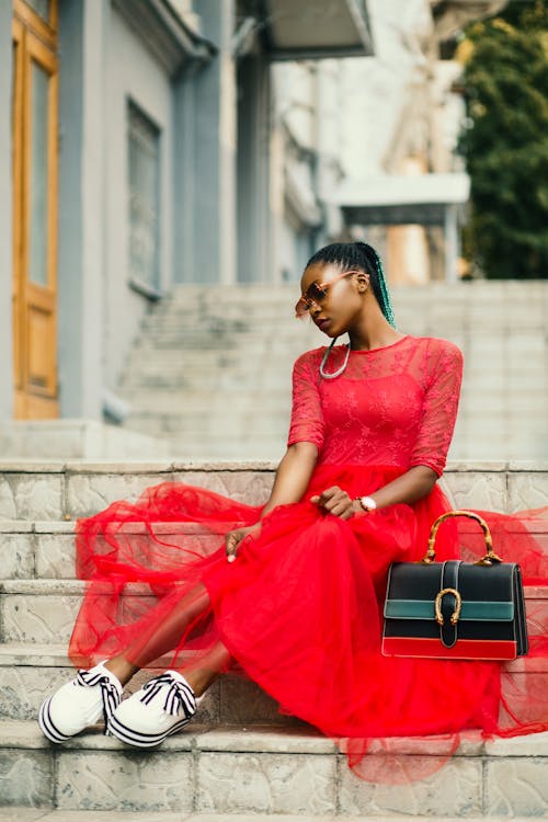 Vrouw Draagt Rode Jurk Met Lange Mouwen In De Buurt Van Zwarte Lederen Tas Zittend Op Betonnen Trap Poseren Voor Fotoshoot
