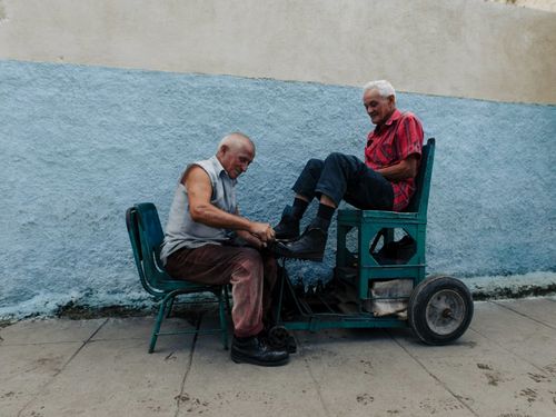 Free Two elderly men interacting outdoors, one cleaning leather shoes of another on a rustic seat. Stock Photo