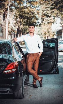 Young man in white shirt confidently posing by open car door outdoors.