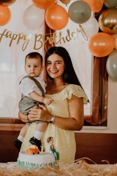 A mother holding her son at a birthday party with balloons and a cake.