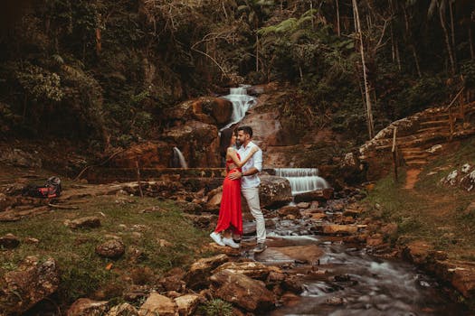 A romantic couple embraces in front of a cascading waterfall surrounded by lush greenery.