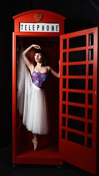 Ballerina in a white tutu posing elegantly inside a red telephone booth.