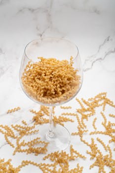 Close-up of uncooked cavatappi pasta in a clear wine glass on a marble surface.