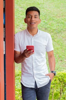 A young man in a white shirt smiling while using a smartphone outdoors.