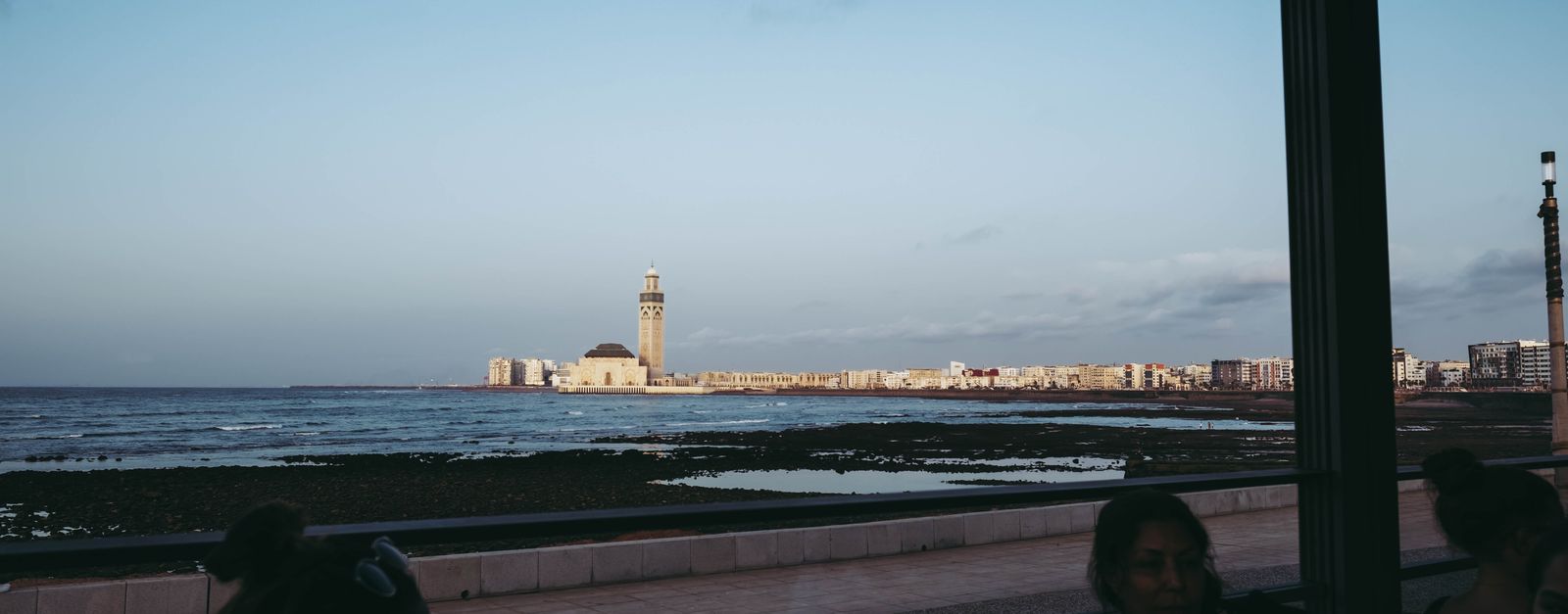 Scenic view of Hassan II Mosque from the Casablanca coastline during daytime with people in a café foreground.