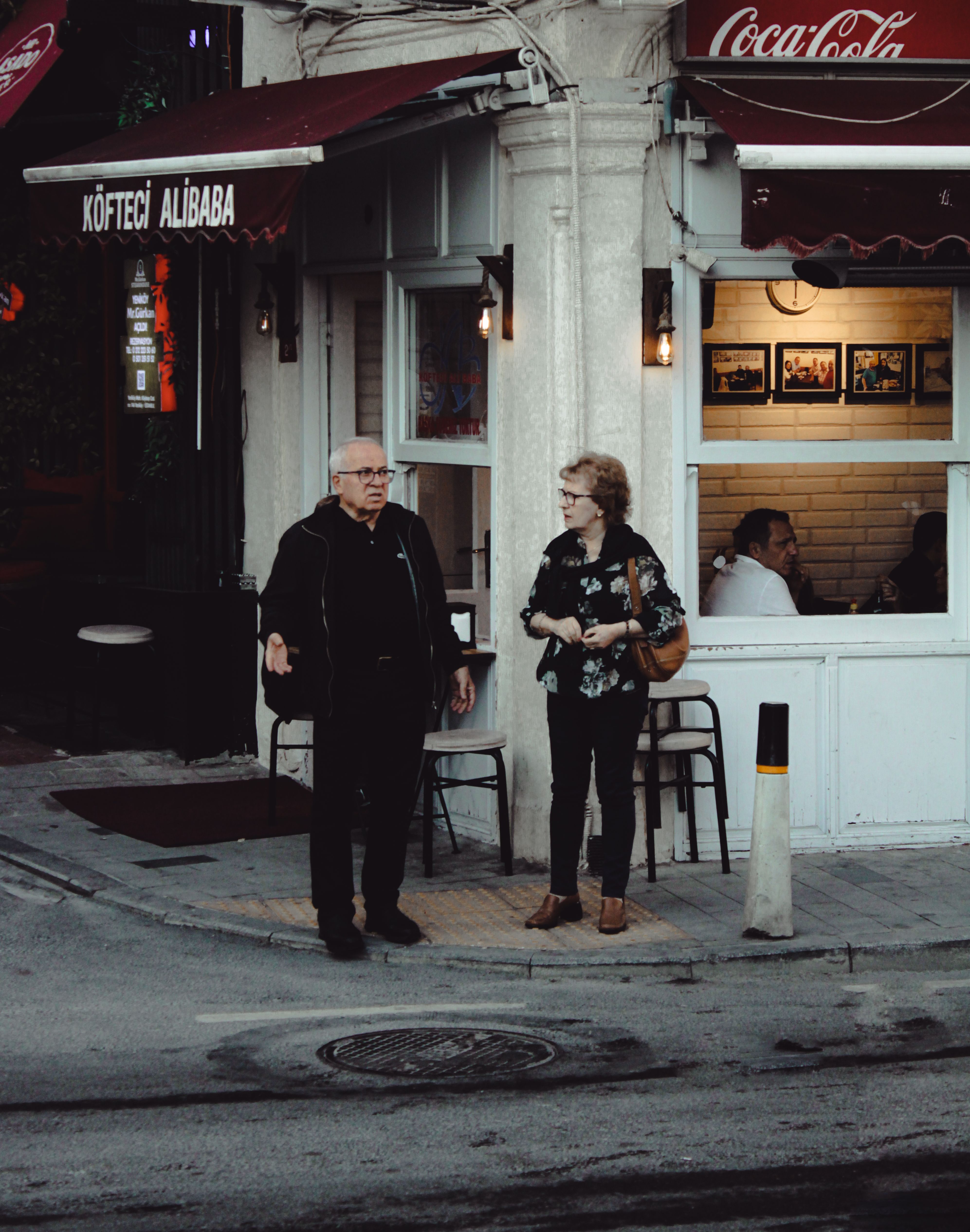 Two people standing outside a small café in the evening, mid-conversation on the pavement