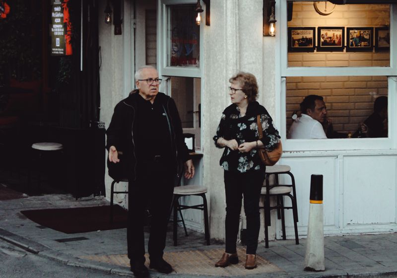 Two people standing outside a small café in the evening, mid-conversation on the pavement